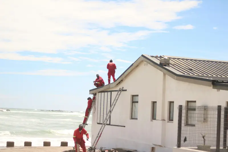 A roofing crew in red overalls on ladders and a metal roof by the sea